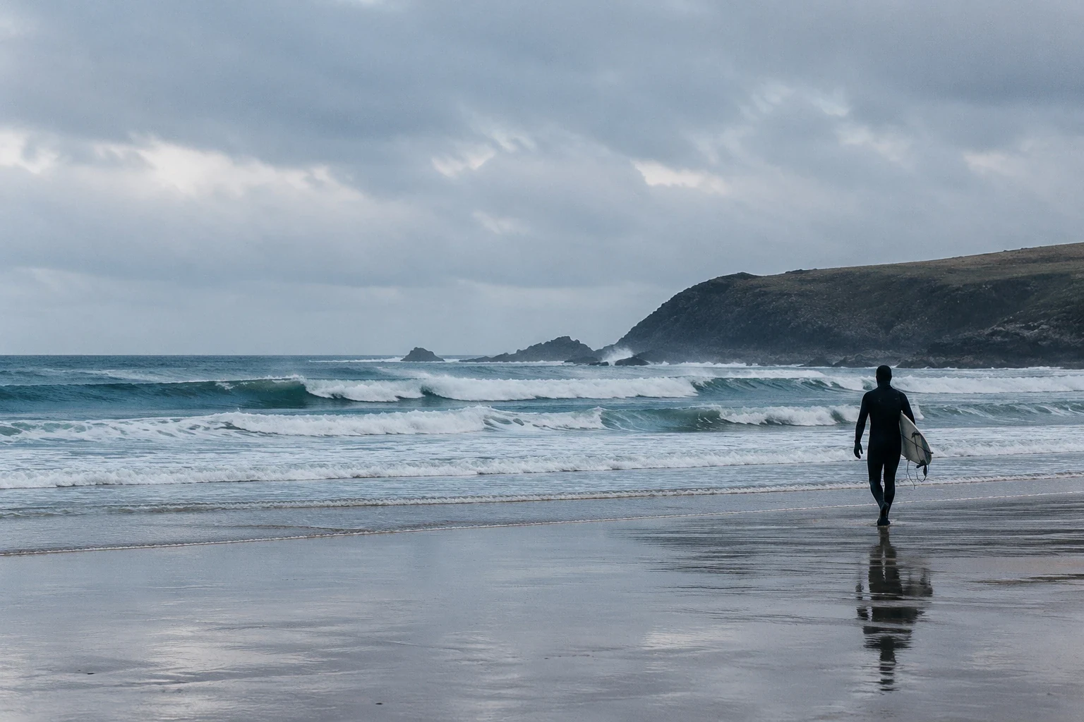 Surfer in a winter wetsuit walking towards grey waves on a Cornish beach